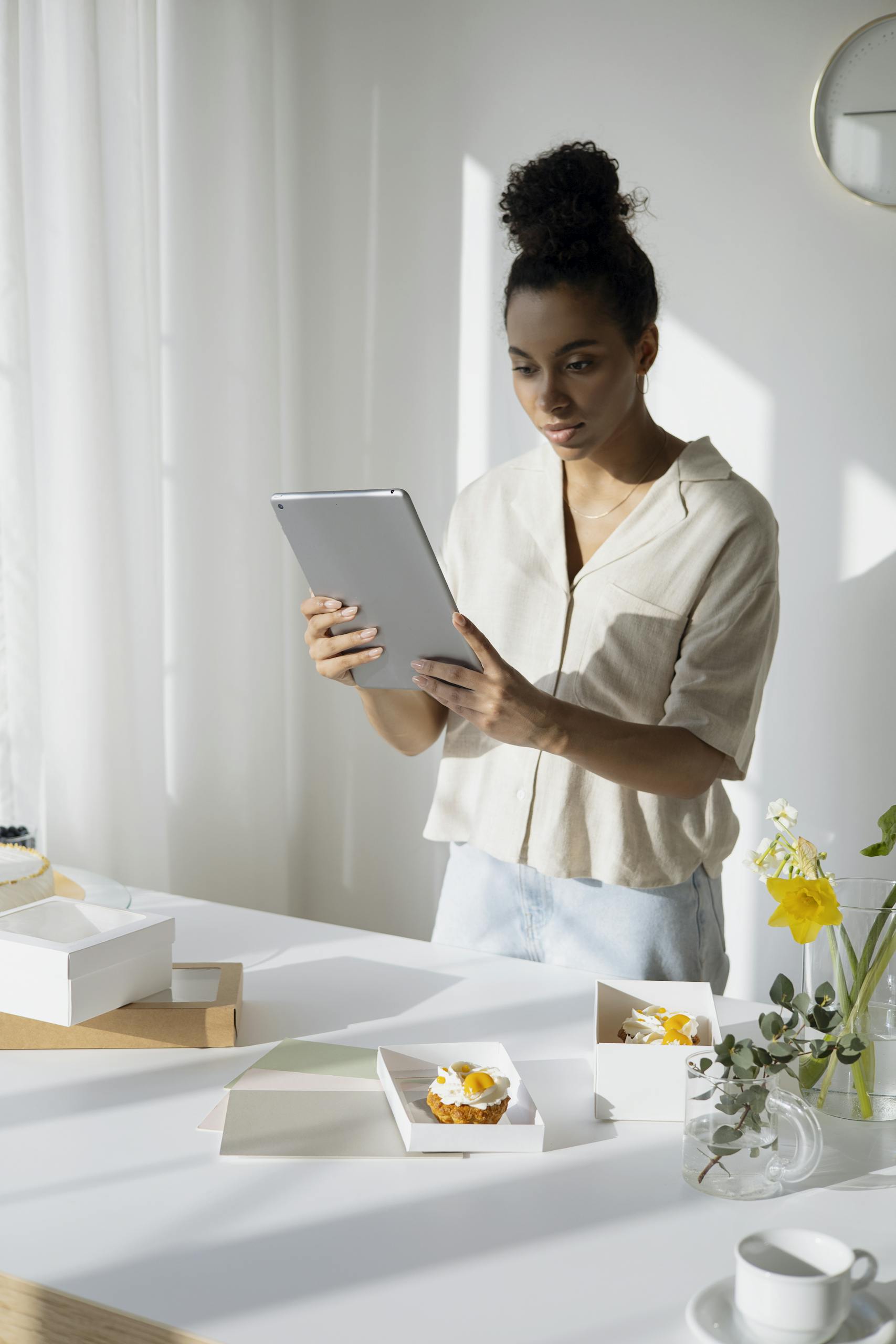 Young woman using a tablet in a bright kitchen, managing her small bakery business and orders.